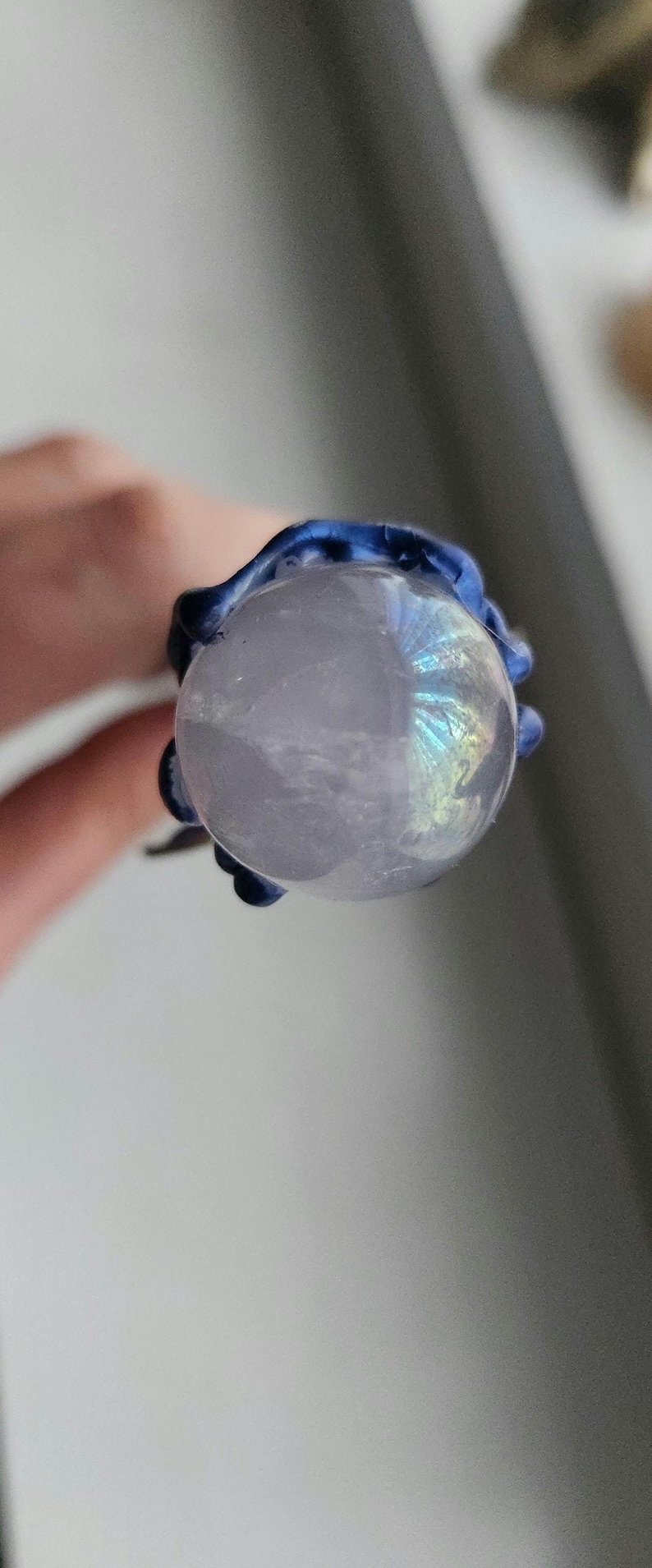 May include: A close-up view of a clear, round, iridescent crystal ball held by a dark blue, textured, claw-like setting. The crystal ball has a subtle, pearly sheen and displays a rainbow of colors. The setting appears to be made of a resin or similar material.
