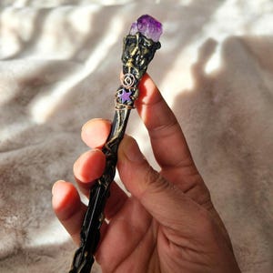 May include: A handcrafted black wand with ornate gold accents, topped with a large, raw amethyst crystal. The wand features a wire-wrapped purple star and a copper-colored spiral design. A unique, fantasy-themed accessory.