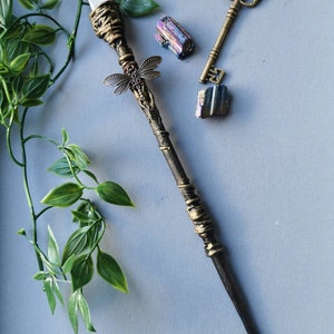 May include: A decorative wand with a clear crystal tip, gold accents, and a dragonfly charm. The wand is on a gray surface with a key and two iridescent crystals.