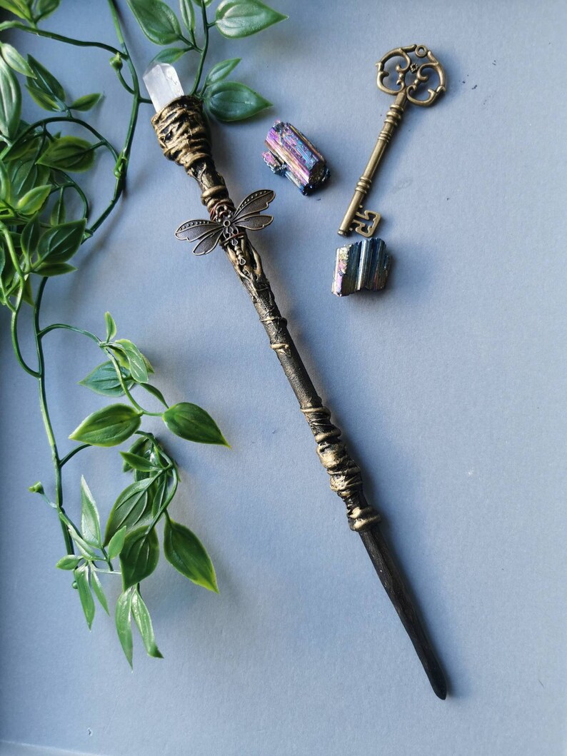 May include: A decorative wand with a clear crystal tip, gold accents, and a dragonfly charm. The wand is on a gray surface with a key and two iridescent crystals.