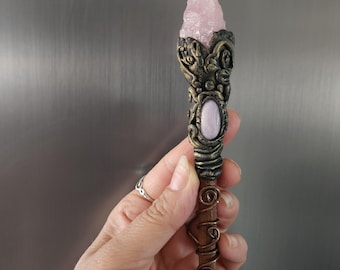May include: A decorative wand featuring a pointed, pale pink raw crystal at the top, set within a dark, textured bronze-colored metal casing. An oval, smooth light pink stone is embedded below the crystal, with copper wire spiraling down the handle.