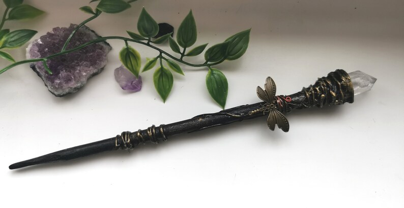 May include: A dark brown wooden wand with a clear quartz crystal tip, wrapped in gold wire and adorned with a bronze dragonfly.