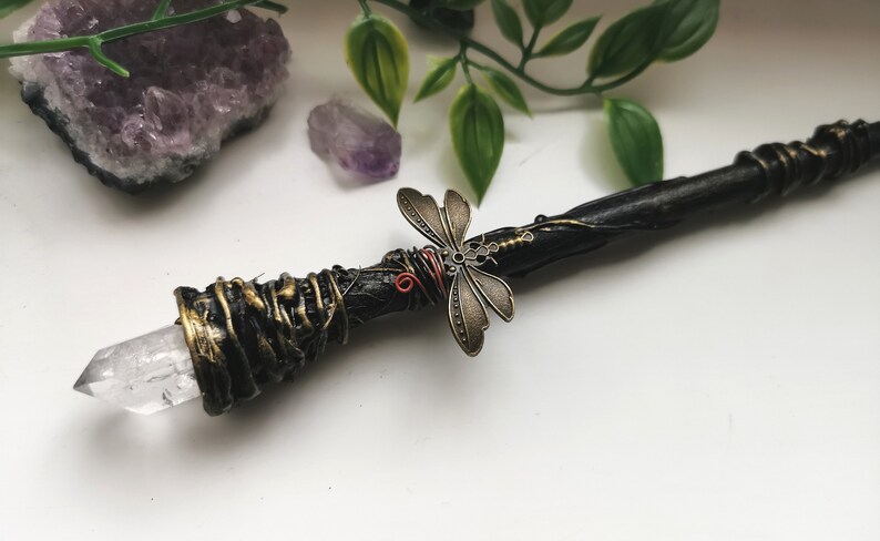 May include: A black and gold wand with a clear crystal tip and a dragonfly charm. The wand is wrapped in gold wire and has a rustic, natural look.