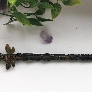 May include: A black wand with a purple amethyst crystal at the tip, wrapped in black wire and decorated with a bronze dragonfly charm.