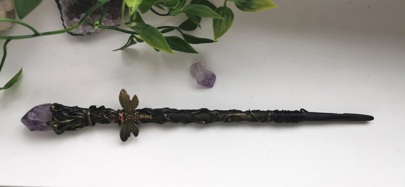 May include: A black wand with a purple amethyst crystal at the tip, wrapped in black wire and decorated with a bronze dragonfly charm.
