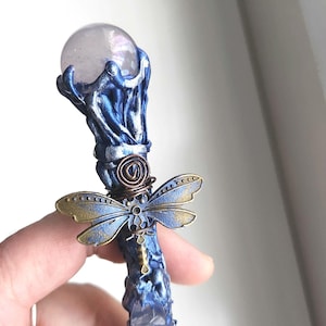 May include: A handcrafted blue and bronze wand with a clear crystal ball at the top. The wand features a dragonfly accent and a crystal embedded in the shaft. The wand is held in a hand.