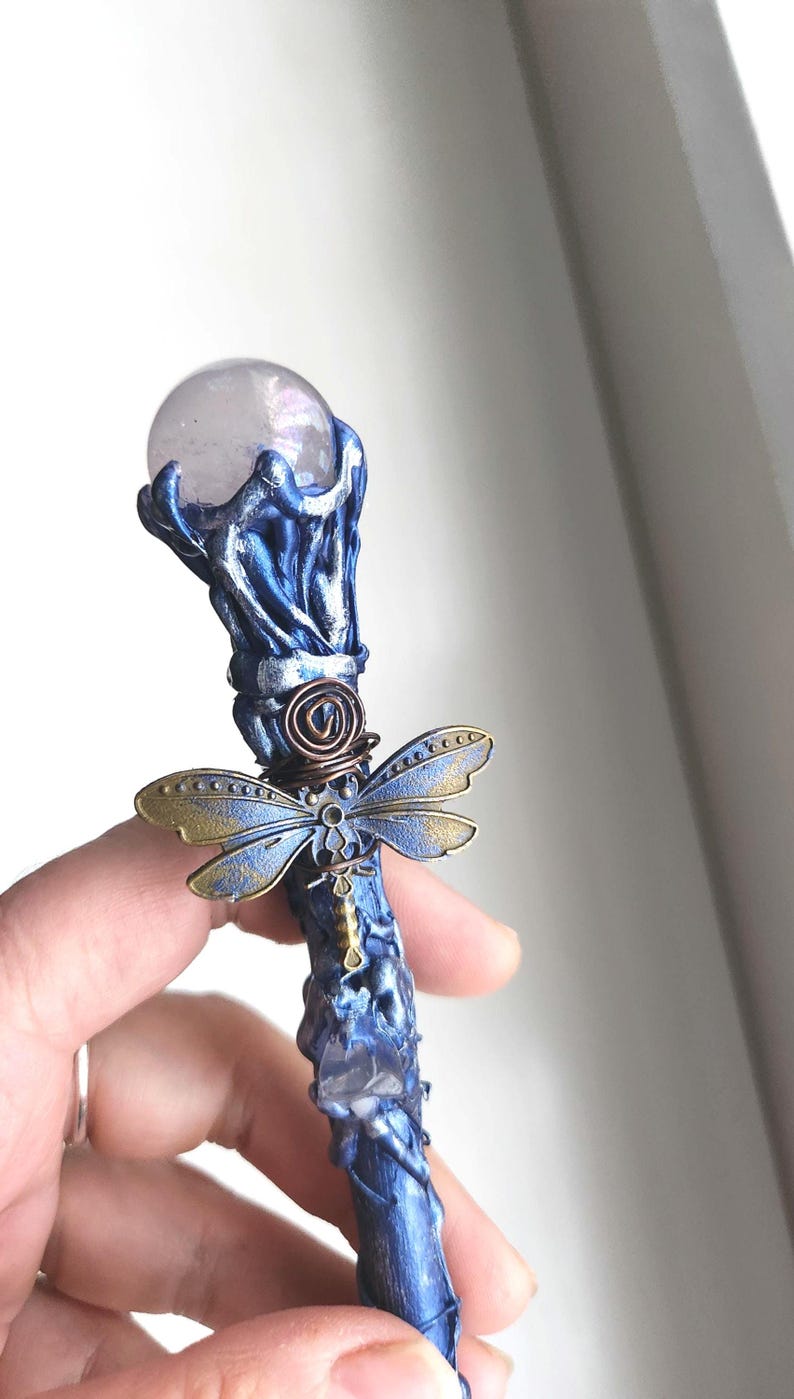 May include: A handcrafted blue and bronze wand with a clear crystal ball at the top. The wand features a dragonfly accent and a crystal embedded in the shaft. The wand is held in a hand.