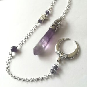 May include: Amethyst crystal pendulum with a silver chain, a crescent moon charm, and a star charm. The crystal is a deep purple colour and has a pointed tip. The chain is silver and has a small bead at the bottom.