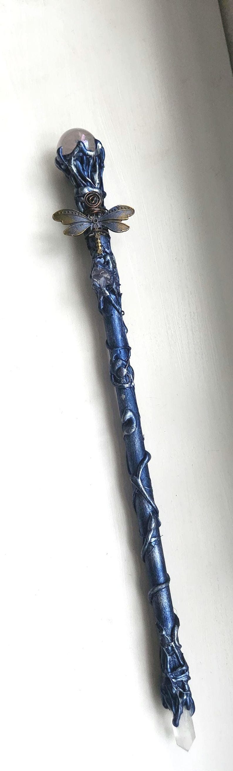 May include: A handcrafted blue and silver wand with a crystal at the base and a crystal ball at the top. A dragonfly charm is affixed to the wand's center. The wand is approximately 12 inches long.