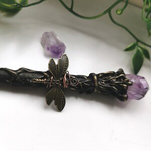May include: A black wand with a purple amethyst crystal at the end. The wand is decorated with a bronze dragonfly and copper wire.