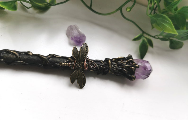 May include: A black wand with a purple amethyst crystal at the end. The wand is decorated with a bronze dragonfly and copper wire.