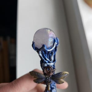 May include: A handcrafted wand with a clear, iridescent crystal ball at the top. The wand features a blue and silver sculpted handle with a bronze dragonfly accent. The wand is approximately 8 inches long.