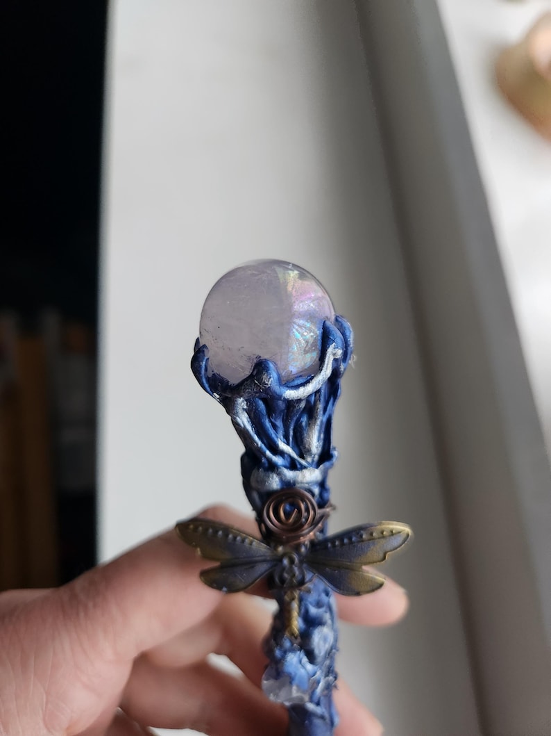 May include: A handcrafted wand with a clear, iridescent crystal ball at the top. The wand features a blue and silver sculpted handle with a bronze dragonfly accent. The wand is approximately 8 inches long.