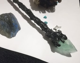 May include: A black wand with a green crystal tip and a silver star charm. The wand is wrapped in black cord and has a rough, textured surface.