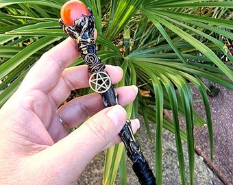 May include: A black and gold wand with a red orb at the top. The wand features a pentagram and detailed metalwork. The wand is held against a backdrop of green foliage.