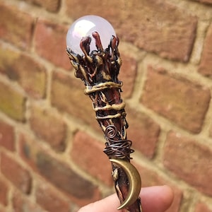 Op de afbeelding: Een handgemaakte houten staf met een heldere kristallen bol aan de bovenkant. De staf heeft ingewikkelde details van brons en koperdraad, een bronzen halve maan en een roodbruine afwerking. De staf wordt in een hand gehouden.