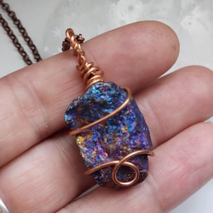 May include: A handcrafted pendant necklace featuring an iridescent stone wrapped in copper wire. The stone displays shades of blue, purple, and gold. The necklace has a copper chain.