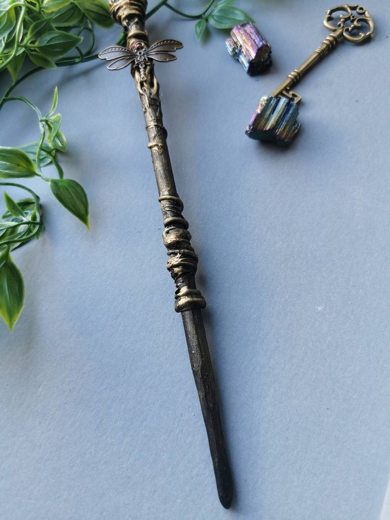 May include: A decorative wand with a dragonfly design at the top. The wand is made of metal and has a dark brown finish. The wand is about 12 inches long.
