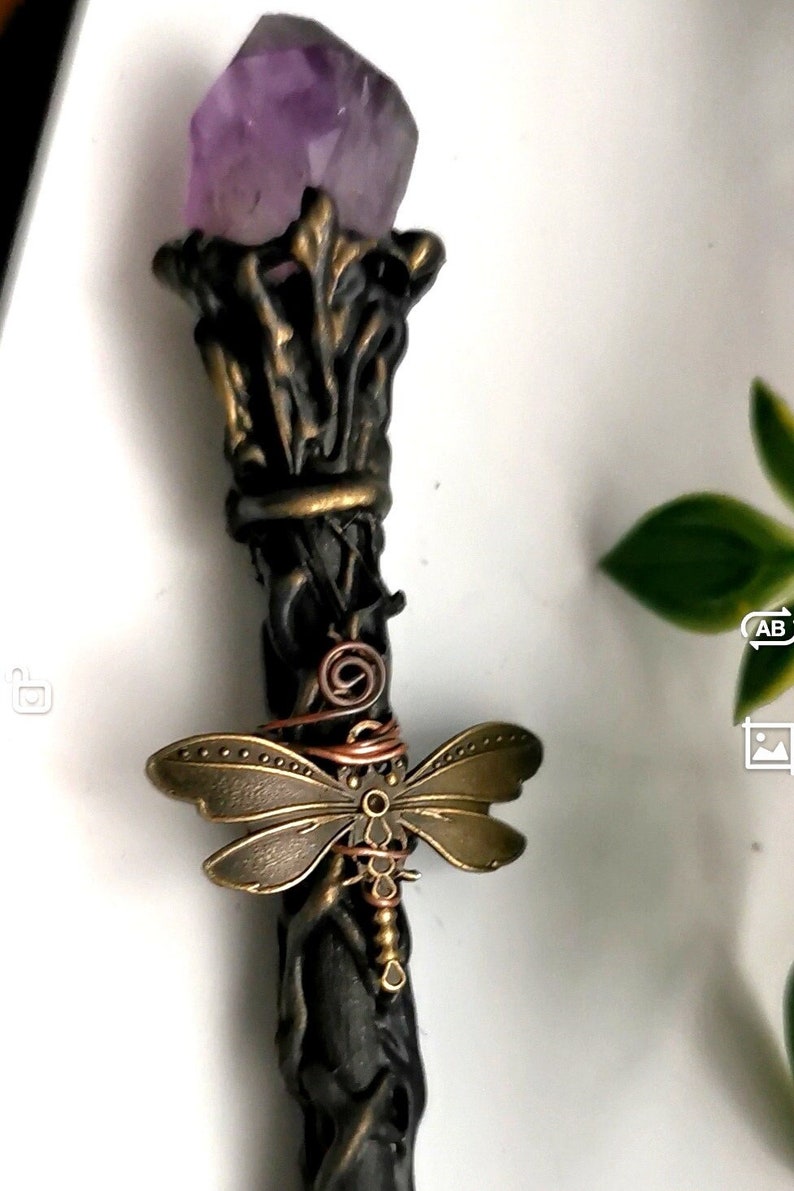 May include: A black wand with a purple amethyst crystal at the top. The wand is decorated with a bronze dragonfly and copper wire.