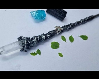 May include: A black and silver wand with a clear quartz crystal tip. The wand has a silver star charm and is surrounded by green leaves.