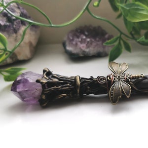 May include: A dark brown wooden wand with a purple amethyst crystal at the tip and a dragonfly charm in the middle. The wand is decorated with intricate details and has a rustic, natural look.