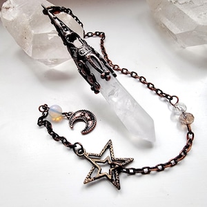 Könnte beinhalten: Ein weißer Quarzkristall-Pendel mit einer Kupferkette und Anhängern. Die Anhänger umfassen einen Halbmond, einen Stern und eine kleine, klare Perle. Der Pendel hängt an einer Kupferkette.