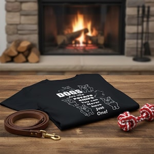 Puede incluir: Camiseta negra con texto blanco e ilustraciones de perros. El texto dice: "¡Los perros son como patatas fritas, nunca puedes tener solo uno!" También hay una correa de cuero marrón y un juguete para perros de cuerda roja y blanca.