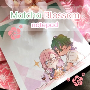May include: A pink and green notepad with the text "Matcha Blossom notepad" and an illustration of two cartoon characters, one with pink hair and glasses and the other with green hair, sitting at a table with a plate of pasta.