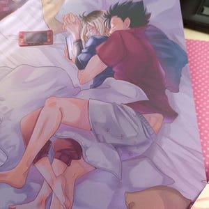 K&ouml;nnte beinhalten: Illustration von zwei Figuren im Bett, die sich umarmen. Eine Figur tr&auml;gt einen weinroten Kapuzenpullover, die andere ein blaues Hemd. Eine Handheld-Konsole und ein Smartphone sind sichtbar. Die Bettw&auml;sche ist wei&szlig;, und die Farbpalette ist weich und warm.