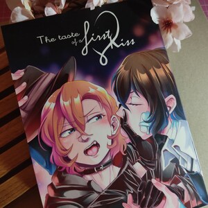 May include: A manga book cover with two characters, one with brown hair and the other with black hair. The title of the book is "The taste of a first kiss".