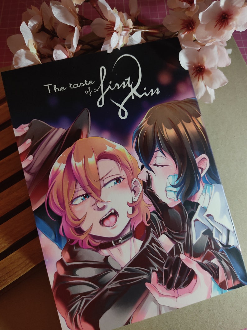 May include: A manga book cover with two characters, one with brown hair and the other with black hair. The title of the book is "The taste of a first kiss".
