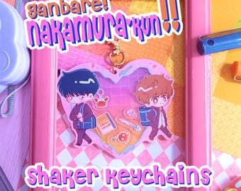 May include: A heart-shaped acrylic shaker keychain featuring two anime-style characters with dark and brown hair, wearing school uniforms and carrying bags. Inside the clear heart are small floating charms including a crab, a book, and pens.