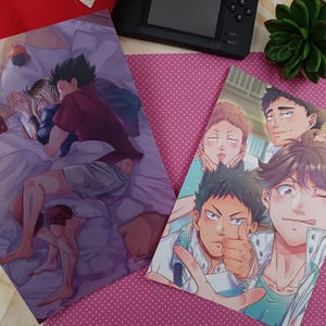 K&ouml;nnte beinhalten: Zwei Anime-Kunstdrucke mit m&auml;nnlichen Charakteren. Einer zeigt zwei Figuren im Bett, der andere zeigt eine Gruppe m&auml;nnlicher Charaktere. Die Drucke befinden sich auf einem rosa gepunkteten Hintergrund mit einer schwarzen Spielkonsole und einer kleinen Pflanze.