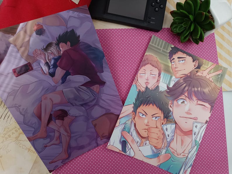 K&ouml;nnte beinhalten: Zwei Anime-Kunstdrucke mit m&auml;nnlichen Charakteren. Einer zeigt zwei Figuren im Bett, der andere zeigt eine Gruppe m&auml;nnlicher Charaktere. Die Drucke befinden sich auf einem rosa gepunkteten Hintergrund mit einer schwarzen Spielkonsole und einer kleinen Pflanze.