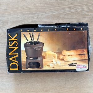 May include: A boxed Dansk fondue set. The box features a dark brown fondue pot with forks, cheese, and bread. The box has the word "Dansk" in large letters on the left side and "Fondue Set" at the top.