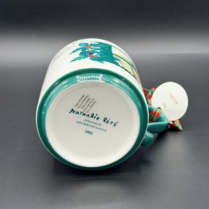 May include: White ceramic mug with a green rim and handle, featuring a colorful illustration. The bottom of the mug displays the text "Nathalie Lete" and "Anthropologie". A tag with the word "From" is attached to the handle.
