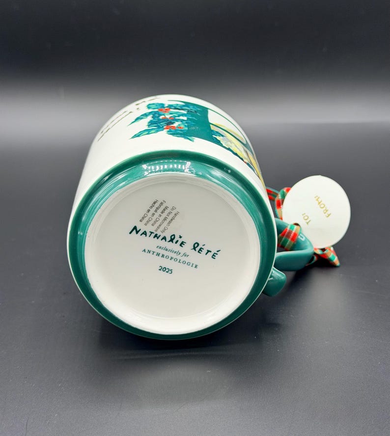 May include: White ceramic mug with a green rim and handle, featuring a colorful illustration. The bottom of the mug displays the text "Nathalie Lete" and "Anthropologie". A tag with the word "From" is attached to the handle.
