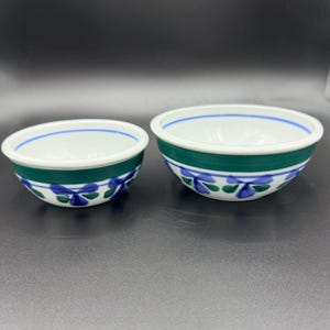 May include: Two ceramic bowls. The bowls have a white interior and a green band around the exterior. Blue floral designs decorate the green band. A blue line accents the rim of each bowl. The bowls are on a dark surface.