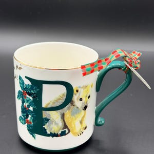 May include: White ceramic mug with a green handle and gold trim. The mug features a large green letter "P" with a polar bear illustration and holly leaves. A plaid ribbon is tied to the handle.