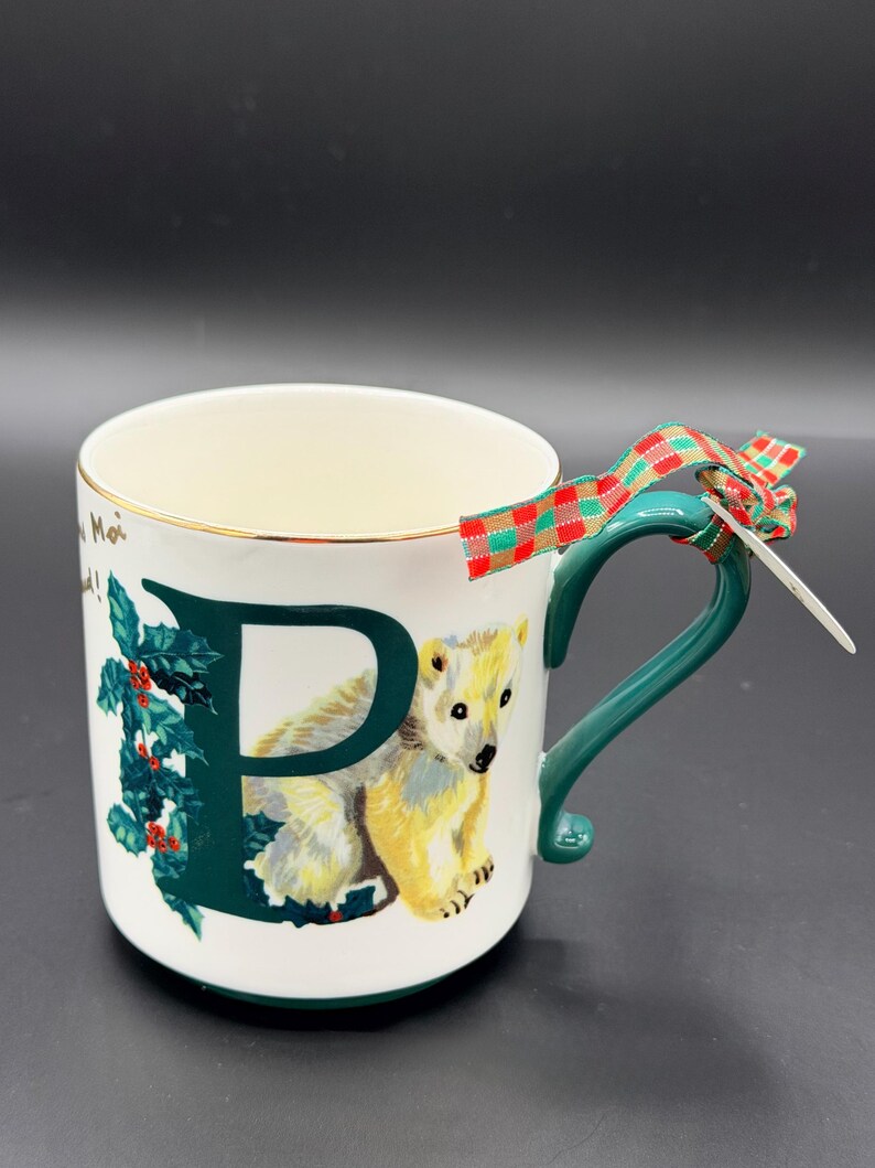 May include: White ceramic mug with a green handle and gold trim. The mug features a large green letter "P" with a polar bear illustration and holly leaves. A plaid ribbon is tied to the handle.