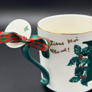 May include: White ceramic mug with a green handle and gold rim. The mug features a holly design and the words "Tiens Moi Chaud!" in gold lettering. A red and green plaid ribbon is tied around the handle with a white tag.