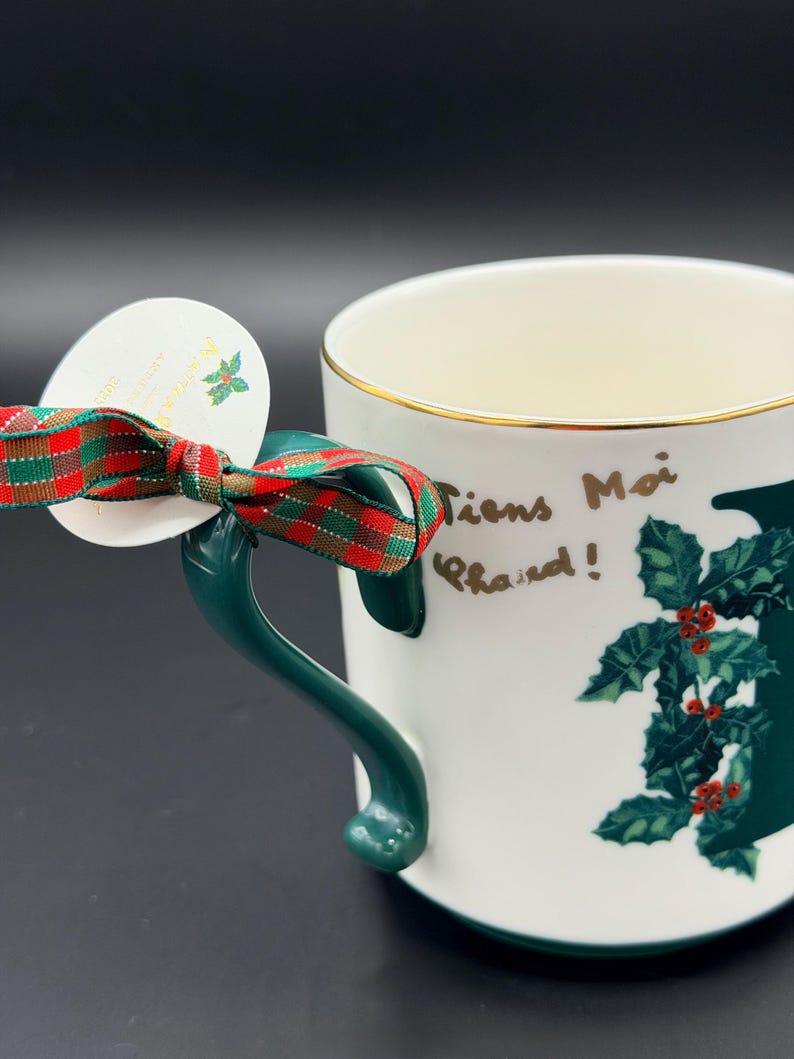 May include: White ceramic mug with a green handle and gold rim. The mug features a holly design and the words "Tiens Moi Chaud!" in gold lettering. A red and green plaid ribbon is tied around the handle with a white tag.