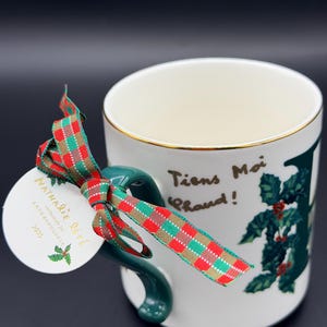 May include: White ceramic mug with a gold rim and a green handle. The mug features a holly design and the French text "Tiens Moi Chaud!" A red, green, and gold plaid ribbon and a tag that reads "Nathalie Lete" are attached to the handle.
