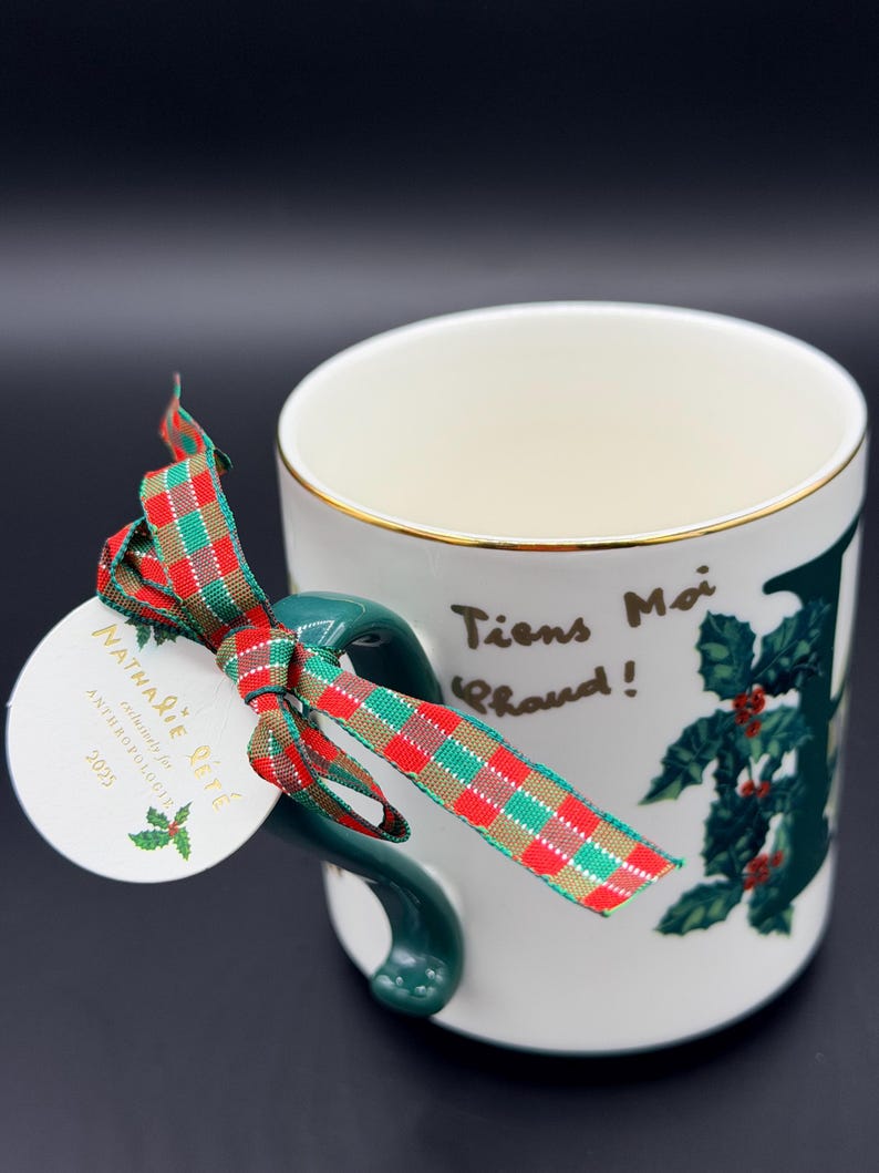 May include: White ceramic mug with a gold rim and a green handle. The mug features a holly design and the French text "Tiens Moi Chaud!" A red, green, and gold plaid ribbon and a tag that reads "Nathalie Lete" are attached to the handle.