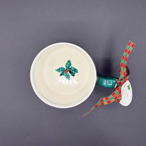 May include: Overhead view of a white ceramic mug with a green handle. The inside of the mug features a holly design with red berries. A red and green plaid ribbon is attached to the handle with a small tag.