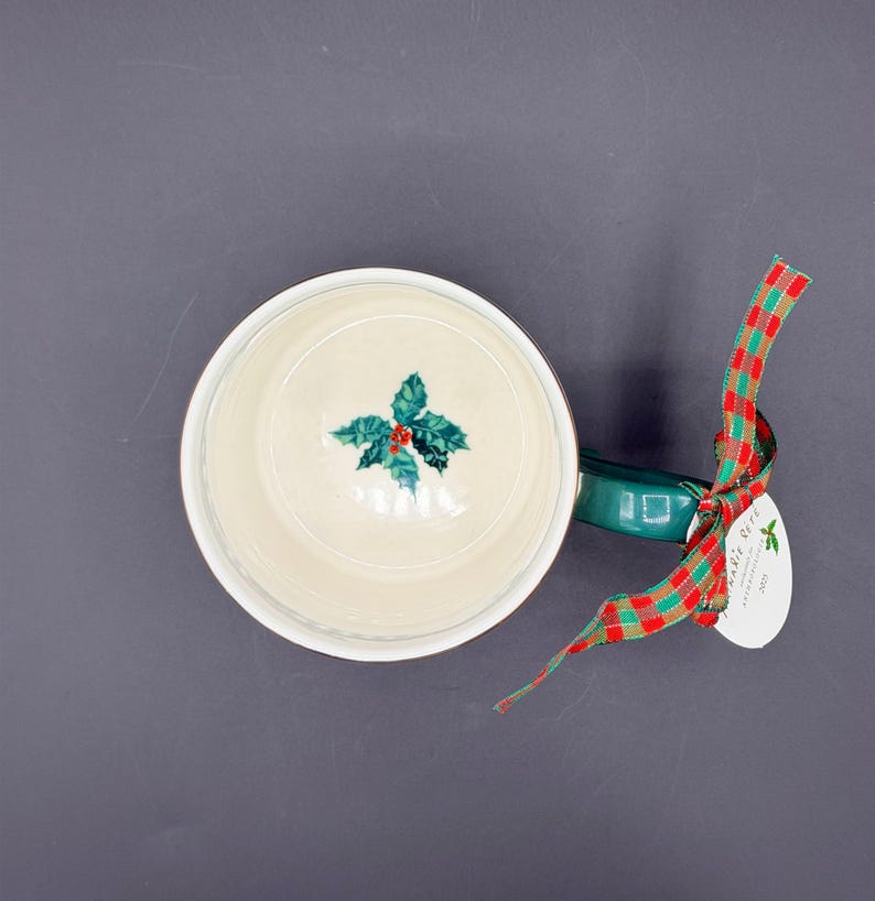 May include: Overhead view of a white ceramic mug with a green handle. The inside of the mug features a holly design with red berries. A red and green plaid ribbon is attached to the handle with a small tag.