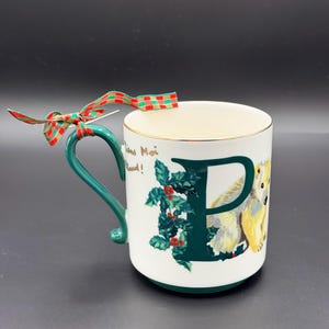 May include: White ceramic mug with a green handle and gold rim. The mug features a large green letter "P" with holly and a polar bear illustration. A red and green plaid ribbon is tied to the handle. The mug has the text "Tiens Moi Chaud!"