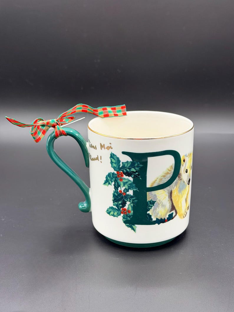 May include: White ceramic mug with a green handle and gold rim. The mug features a large green letter "P" with holly and a polar bear illustration. A red and green plaid ribbon is tied to the handle. The mug has the text "Tiens Moi Chaud!"