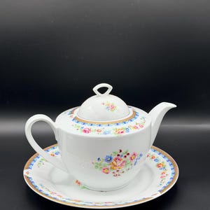 May include: White porcelain teapot and saucer set. The teapot has a lid and a curved handle. Both the teapot and saucer are decorated with a floral pattern in pink, blue, and yellow. The rims are trimmed with gold.