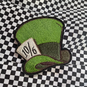 Può includere: Toppa di tessuto ricamata verde e marrone di un cappello con una carta da gioco che dice "10/6" attaccata al lato.